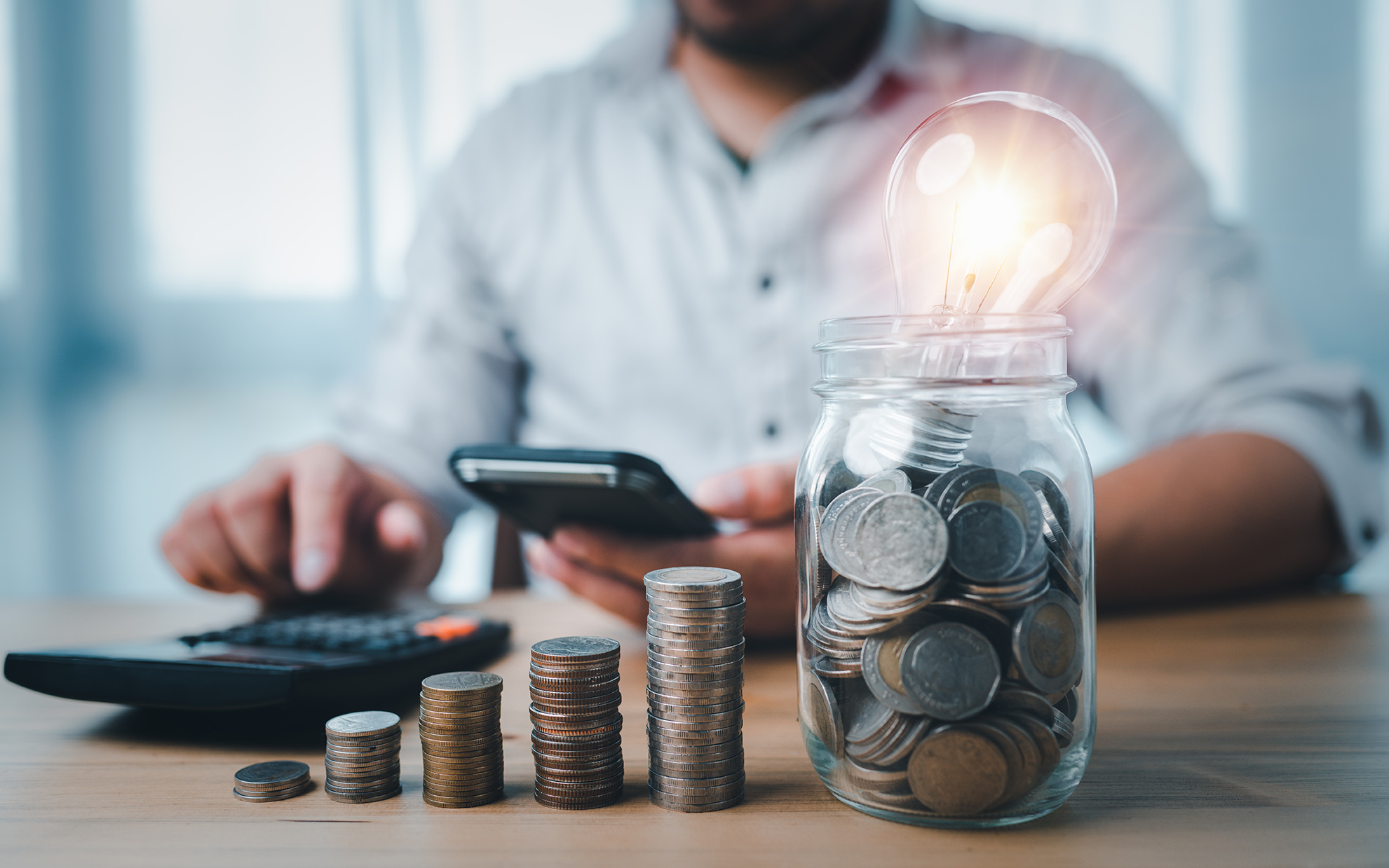 A businessman at a desk with stacked coins and a lightbulb in a jar